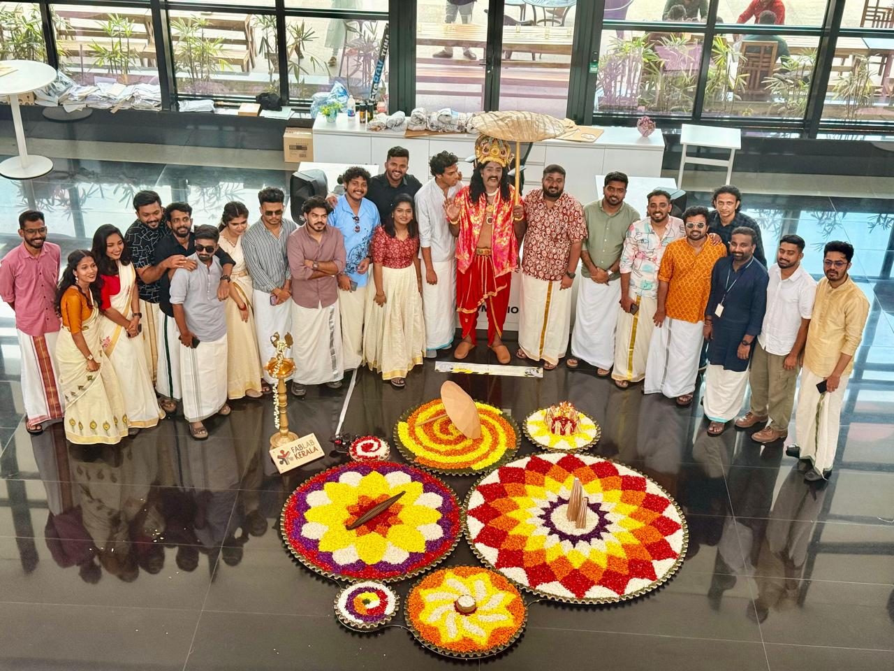 Startup teams standing around finished pookalams laid out across the atrium floor, with Mahabali in costume